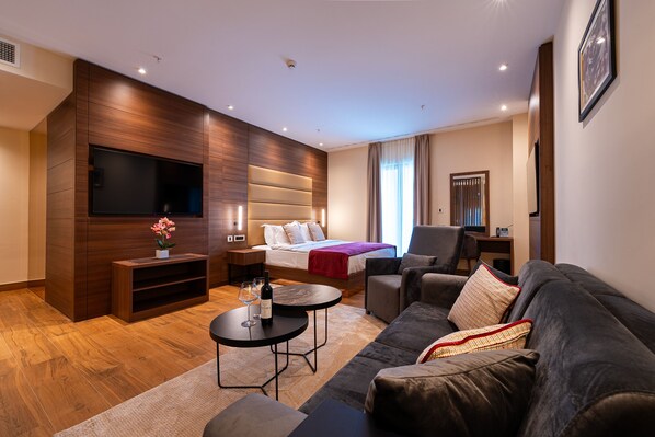 Una habitación de hotel moderna con un televisor de pantalla plana grande, una cama con manta roja, un sofá, una mesita de café y un pequeño mueble de televisión con una copa de vino.