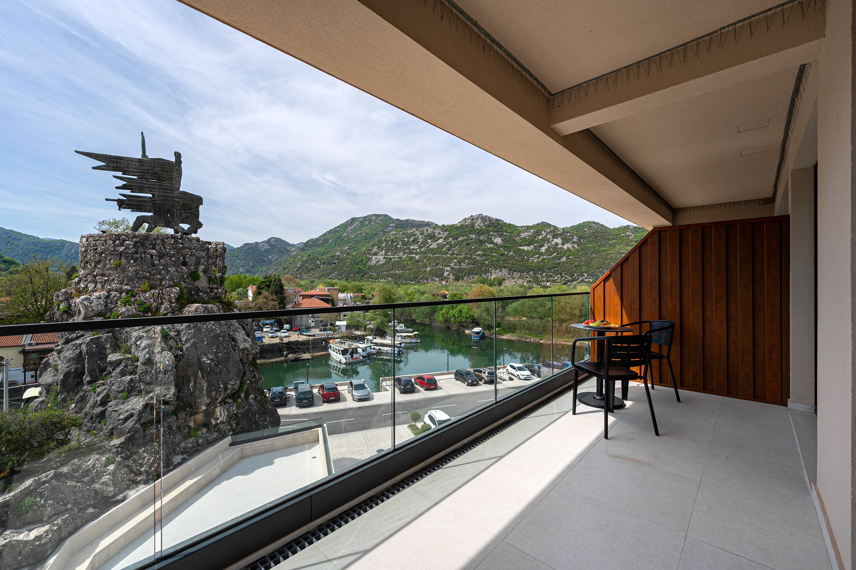 A balcony with a view of a marina, a statue, and a mountainous landscape.