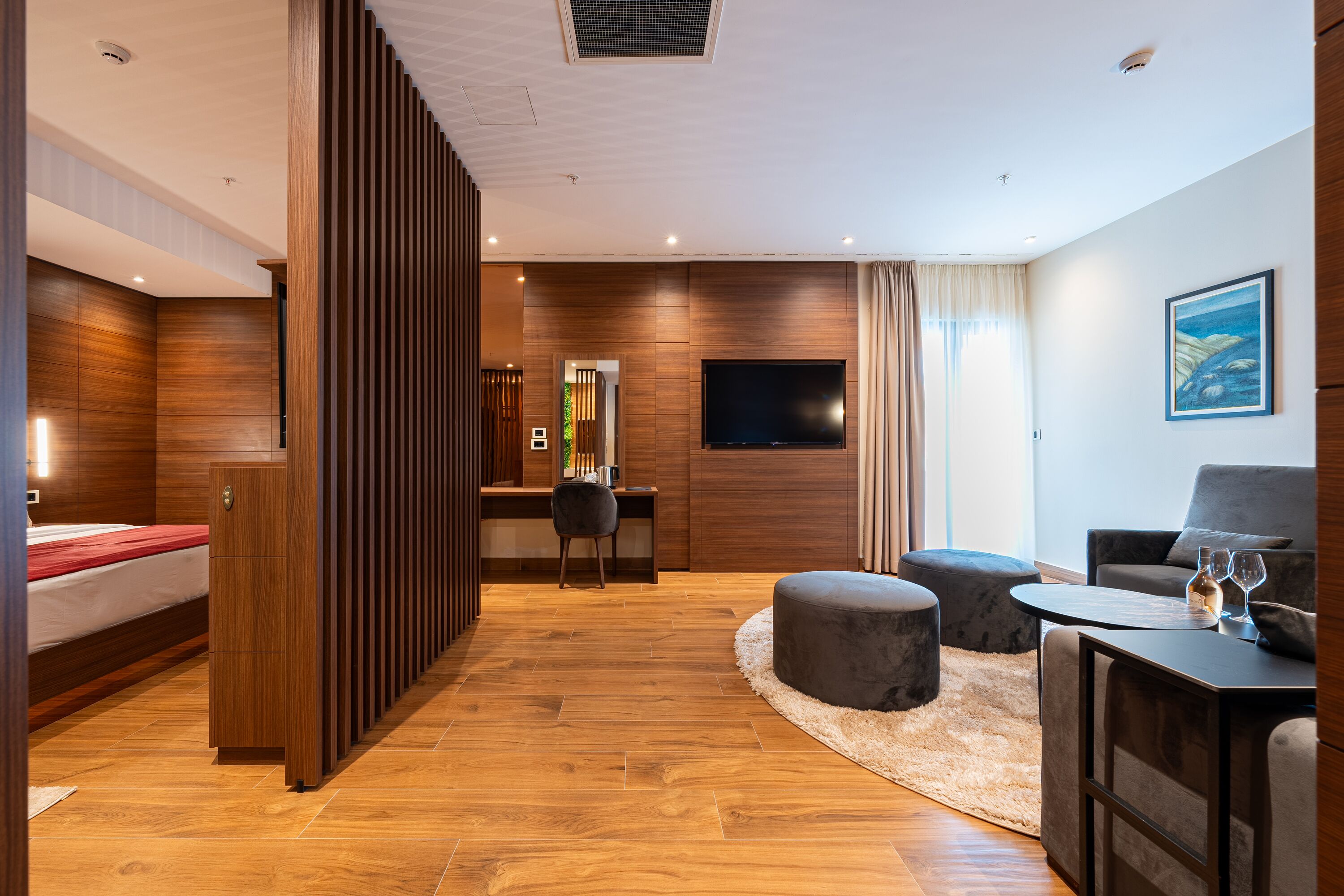Une chambre d’hôtel moderne avec un plancher en bois, un lit, un bureau avec une chaise, une télévision et un coin salon avec des poufs et une table basse.
