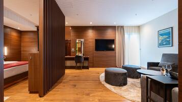 Une chambre d’hôtel moderne avec un plancher en bois, un lit, un bureau avec une chaise, une télévision et un coin salon avec des poufs et une table basse.