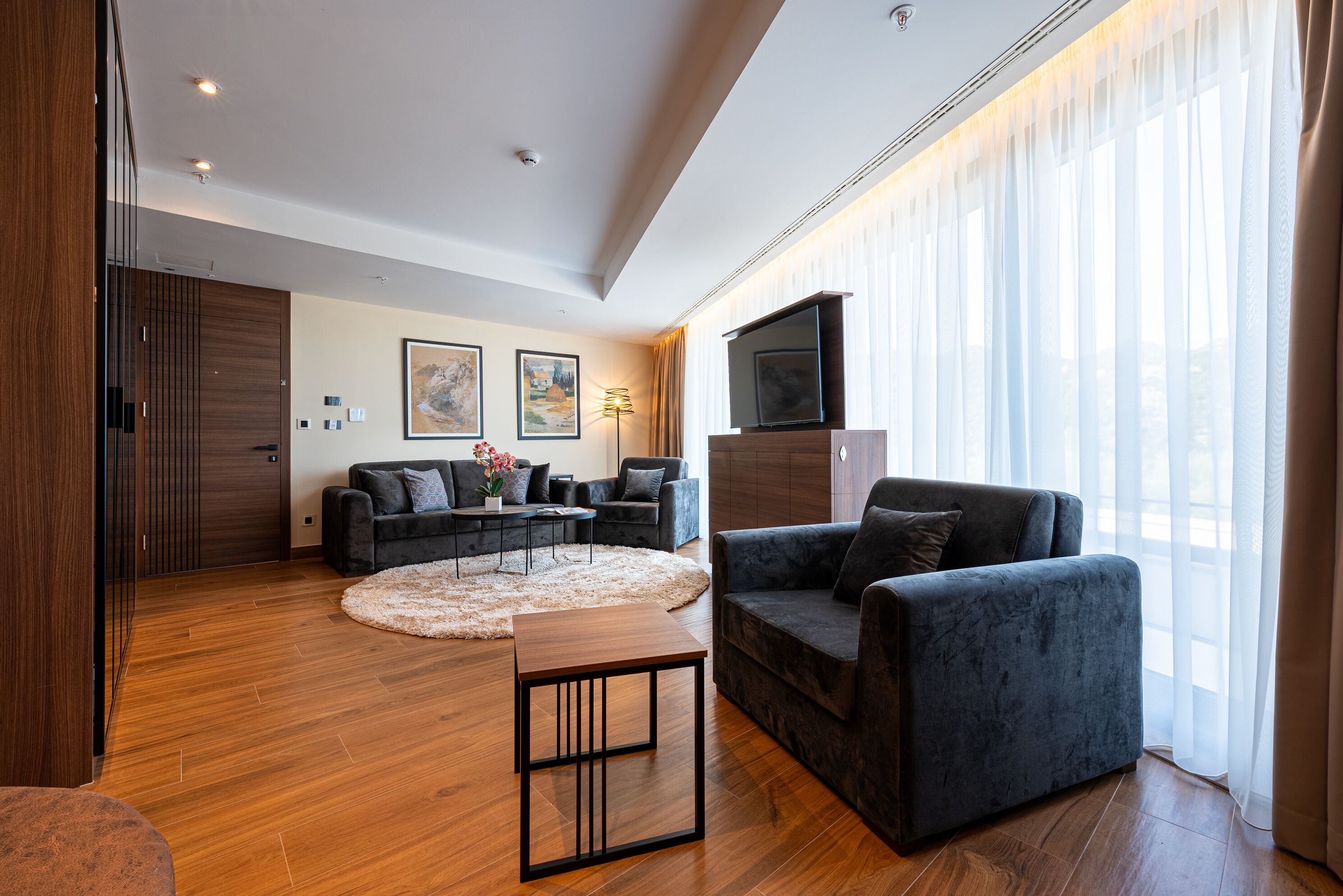 Una sala moderna con sofá, sillón y mesa de centro, piso de madera y un ventanal con cortinas traslúcidas.