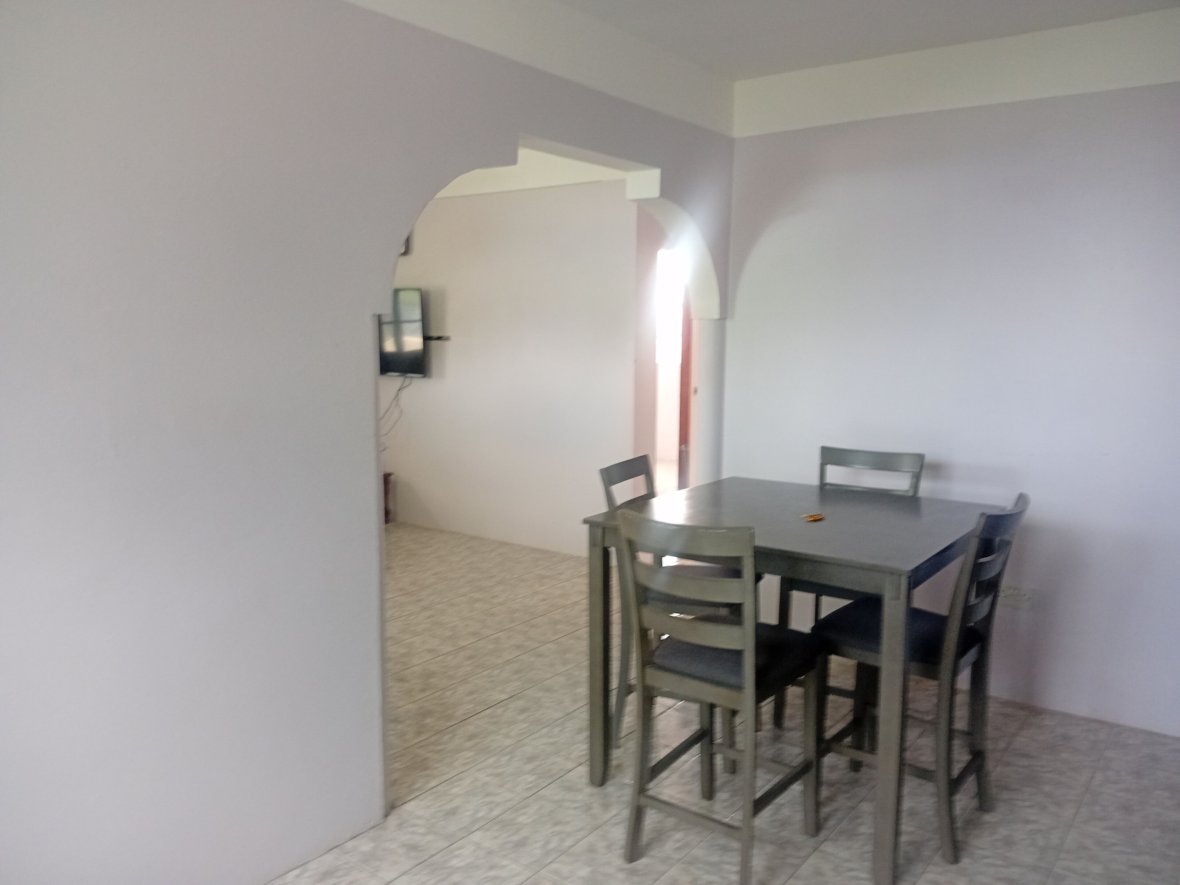 Un comedor con una mesa y cuatro sillas, un televisor de pantalla plana montado en la pared y piso de baldosas.