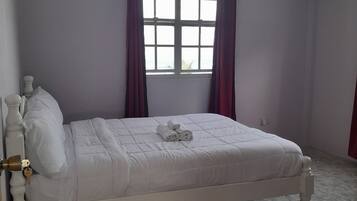 A neatly made bed with a white headboard, a window with purple curtains, and a ceiling fan.