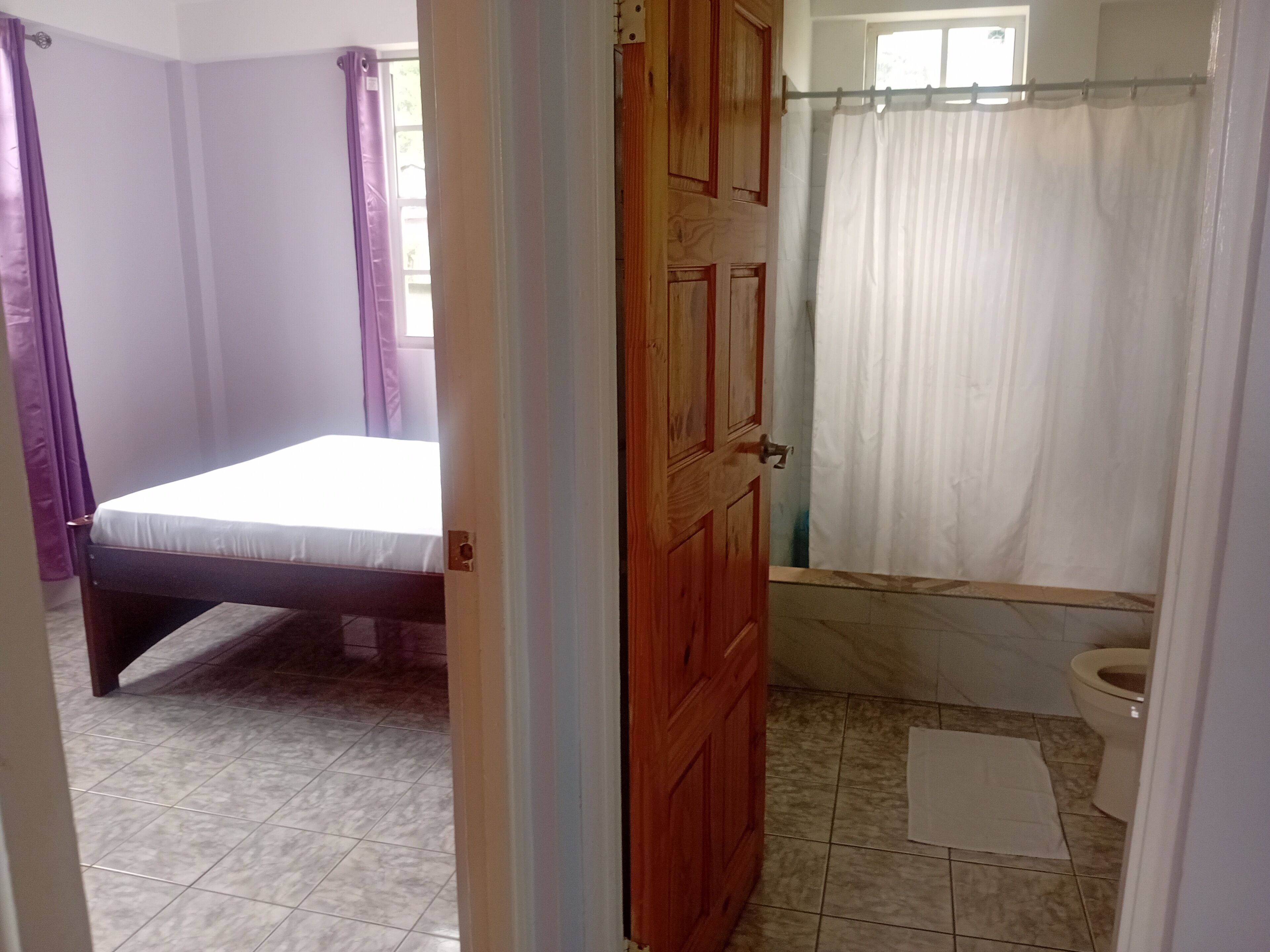 A room with a bed, a wooden door, a shower area with a curtain, and a small white rug.