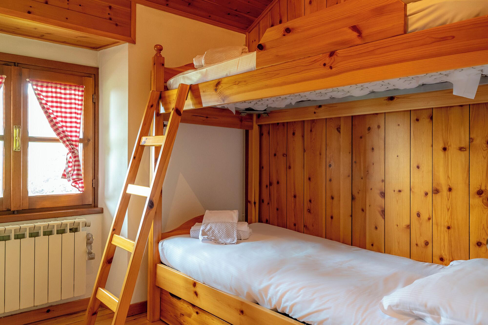 A bunk bed with a wooden ladder, a window with checkered curtains, and a radiator.