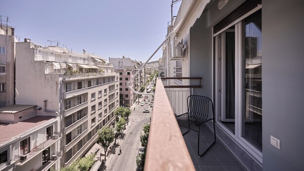 A balcony with a view of a city street, a chair, and a window with a curtain. - Vision Stournari 65 (Athens)