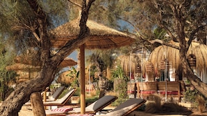 Un área de estar al aire libre tranquila con sillones de sol y un paraguas de paja, rodeada de árboles y un edificio rústico al fondo.