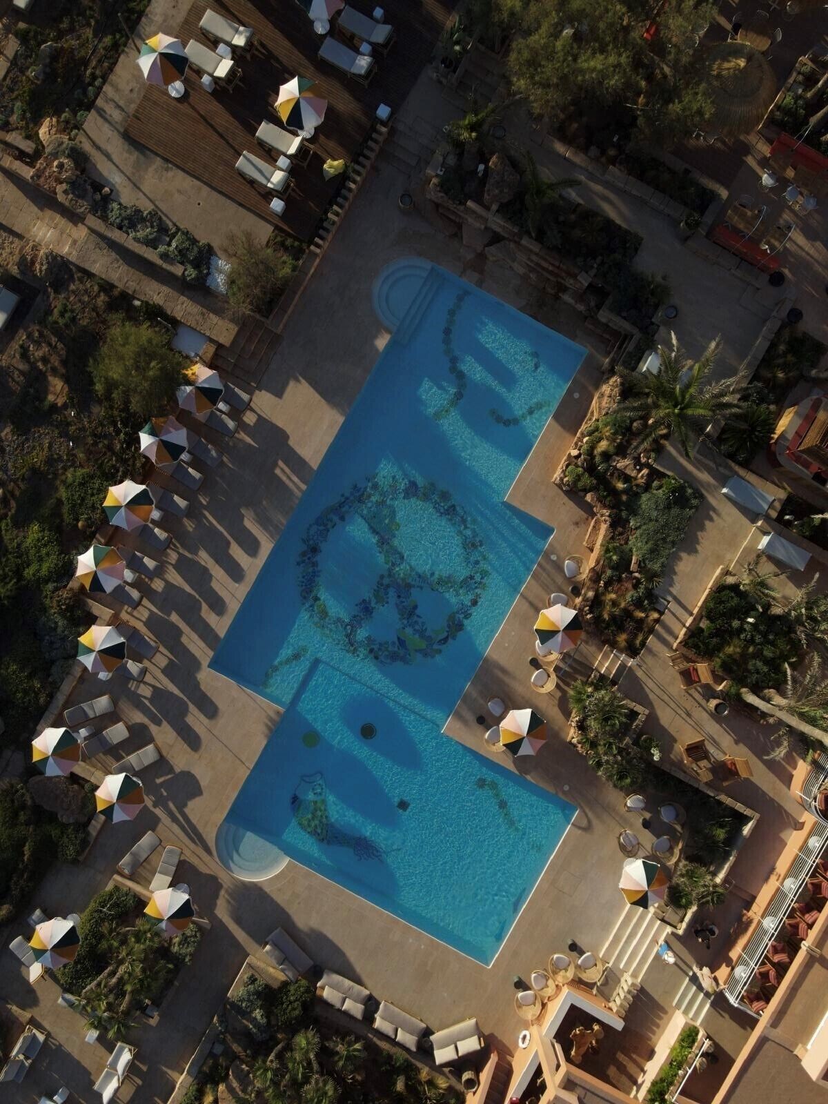 Vista aérea de un resort con una amplia piscina rectangular, tumbonas y sombrillas.