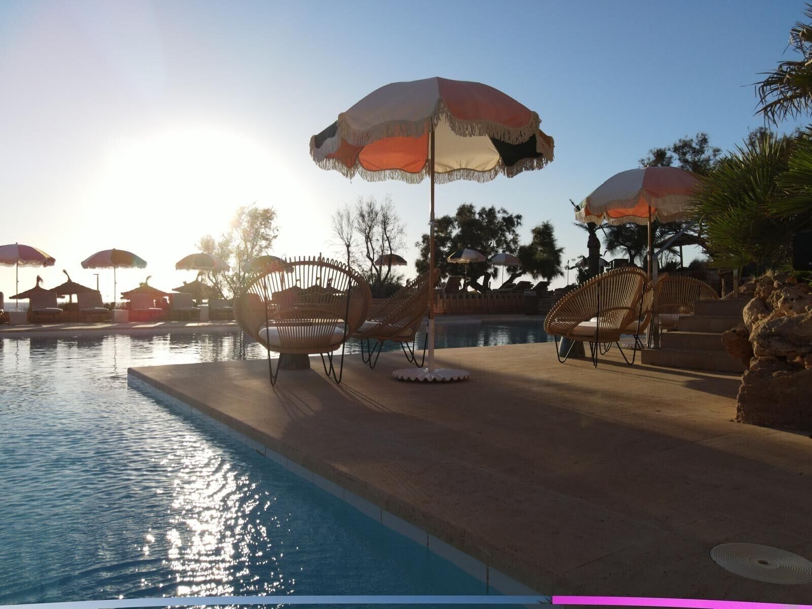 A poolside area with lounge chairs and umbrellas, surrounded by trees and a clear sky.
