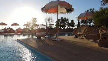 A poolside area with lounge chairs and umbrellas, surrounded by trees and a clear sky.