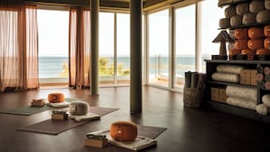Una habitación amplia con ventanas grandes que ofrecen vistas al mar, un espacio para yoga y un estante con almohadas apiladas ordenadamente y esterillas enrolladas.