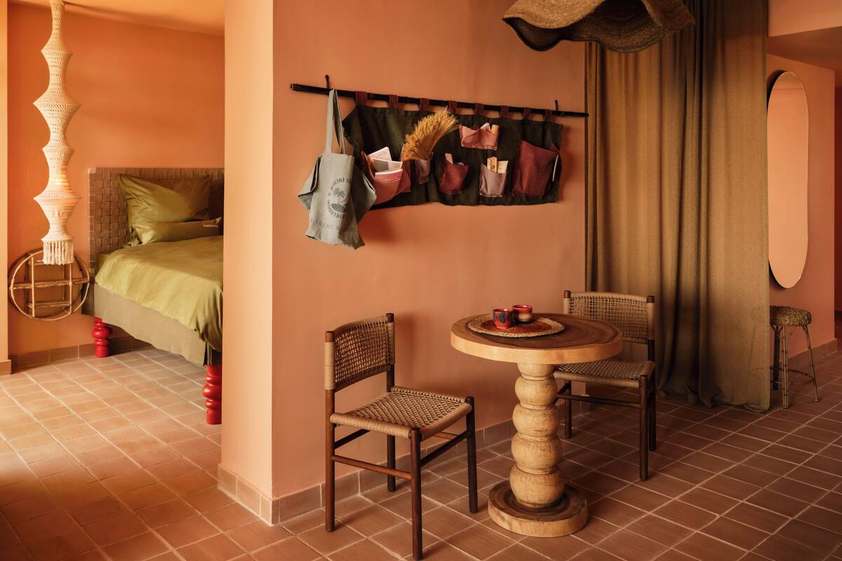 Une chambre d’hôtel confortable avec un lit, une table ronde en bois entourée de chaises, un luminaire suspendu et un porte-manteau fixé au mur, avec des sacs.