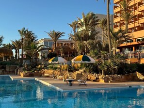 Ein Poolbereich mit Liegestühlen und Sonnenschirmen, umgeben von Palmen und einem Gebäude mit Balkonen.