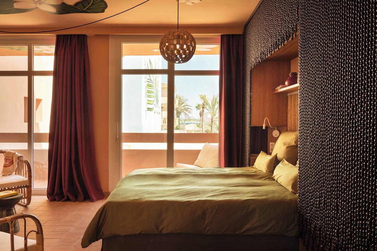 Une chambre d’hôtel avec un grand lit, une chaise, une table et une vue sur les palmiers par la fenêtre.