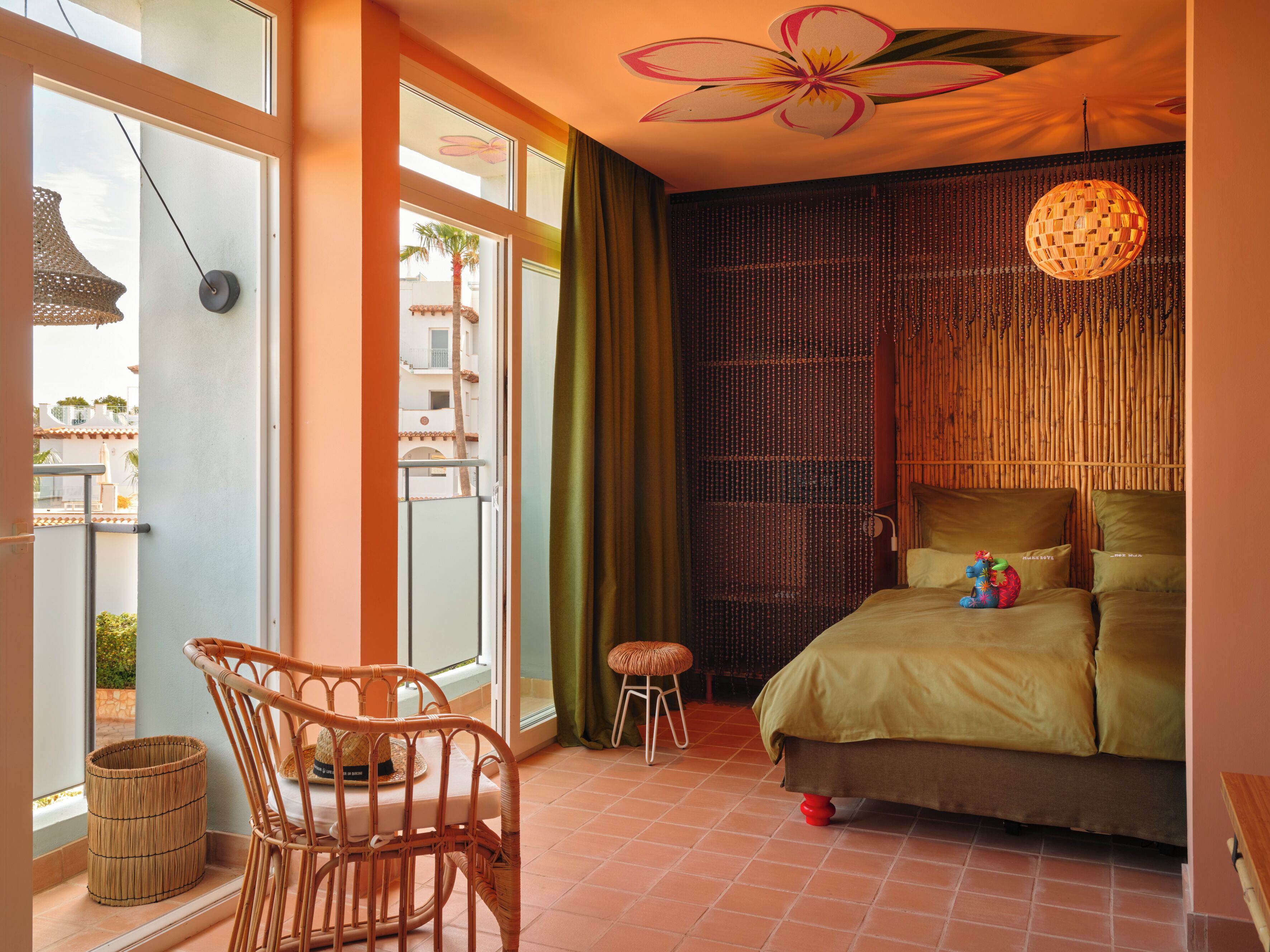 Un dormitorio con cama, una silla, un taburete pequeño, una cesta de mimbre y vistas al balcón exterior.
