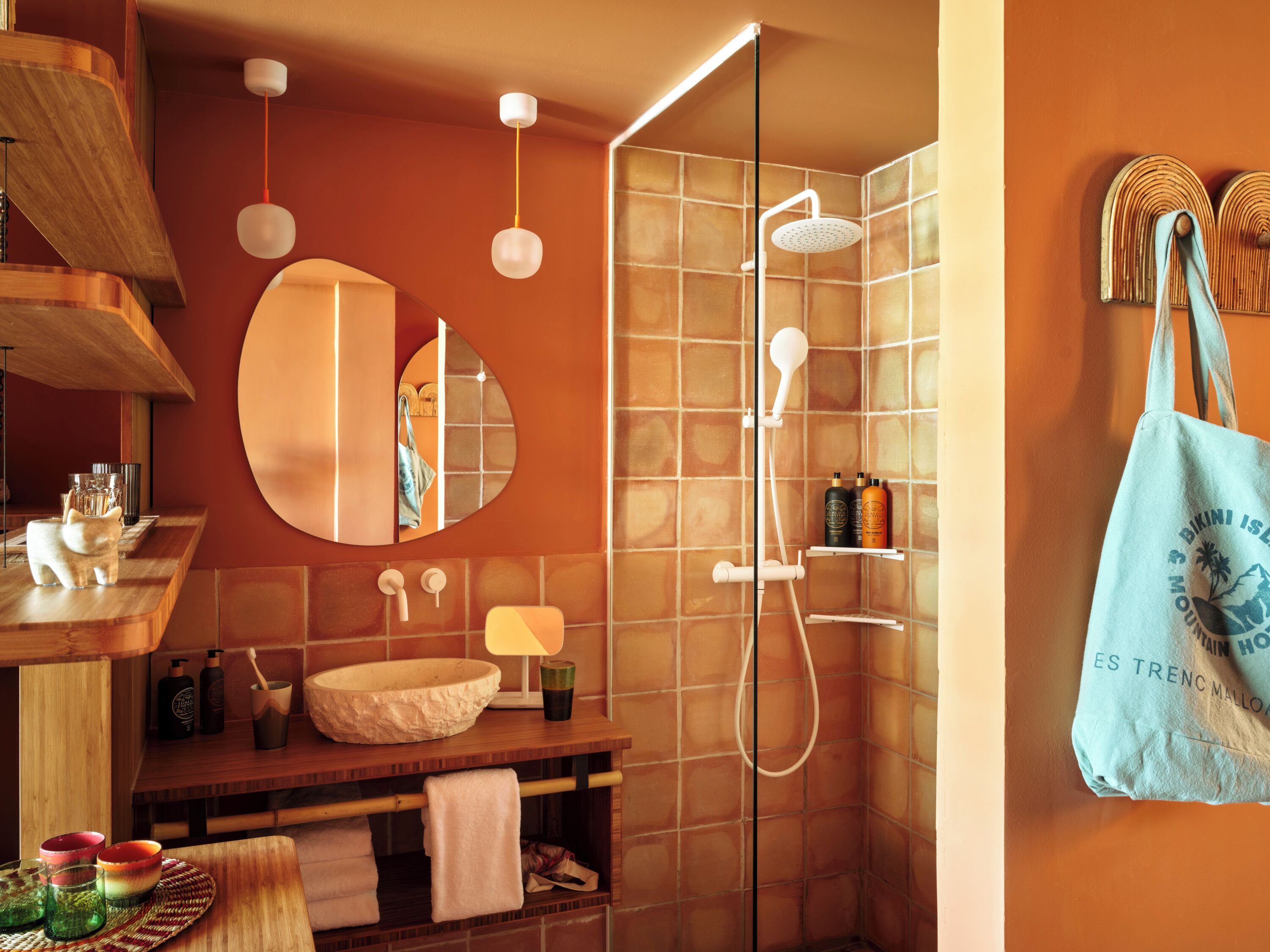 Une salle de bain avec un meuble-lavabo en bois, un miroir rond, une cabine de douche en verre et un porte-serviettes avec un sac bleu.