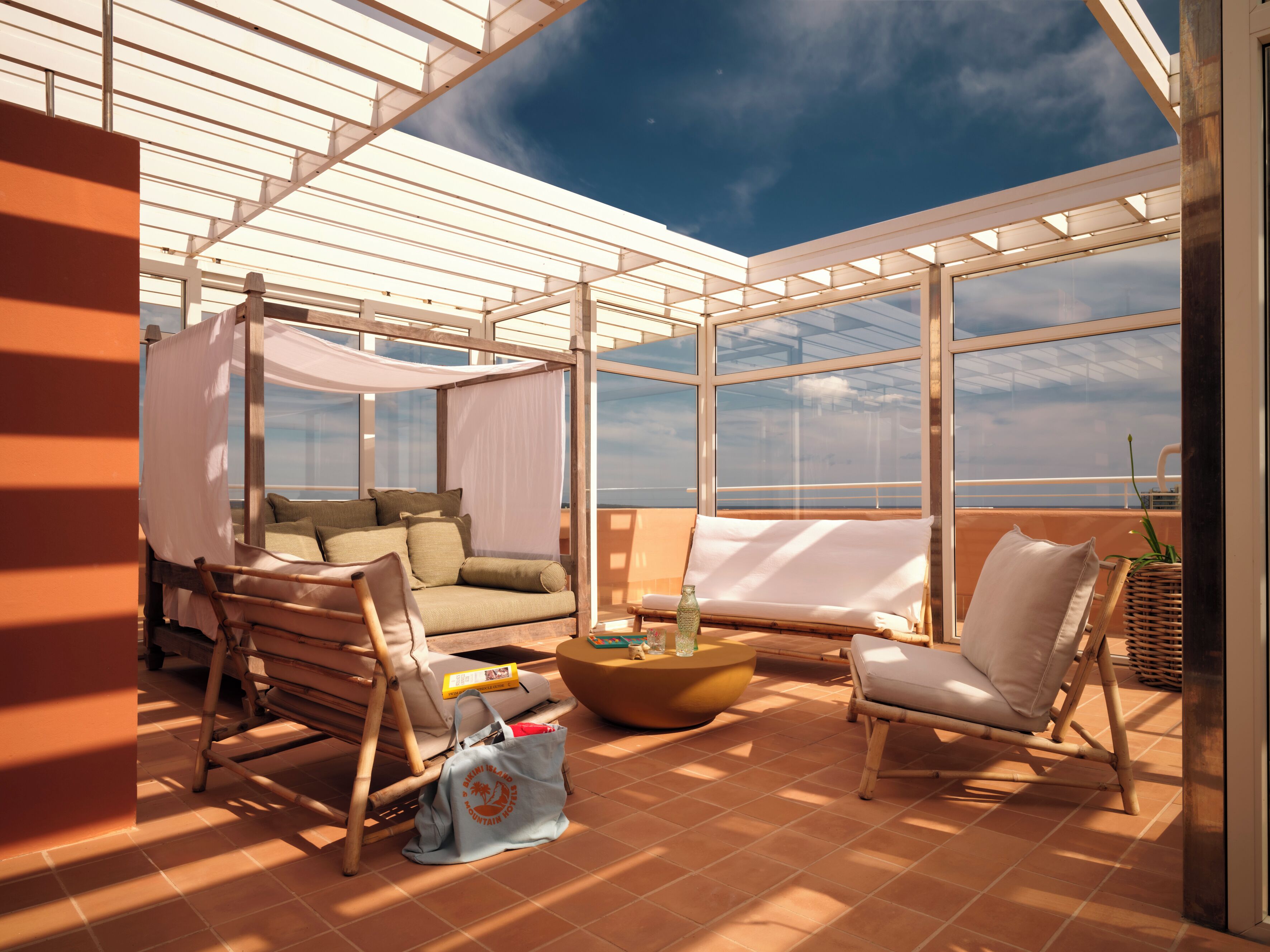 Un espacio al aire libre con luz solar y vistas al océano, con una silla acolchada, una mesita y una silla de mimbre.