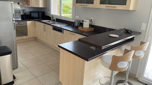 A modern kitchen with light wood cabinets, a stainless steel refrigerator, a built-in oven, a black countertop, and a window with a view of greenery. - Maison D'hôtes (Baslieux-sous-Châtillon)