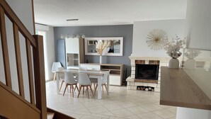 A modern living room with a dining area, a fireplace, and a staircase. - Maison D'hôtes (Baslieux-sous-Châtillon)