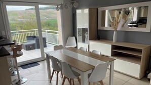 A modern kitchen with a wooden table and white chairs, a built-in oven, and a large window overlooking a green landscape. - Maison D'hôtes (Baslieux-sous-Châtillon)