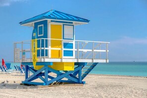 A lifeguard tower with a blue and yellow color scheme, located on a sandy beach with clear blue water in the background.