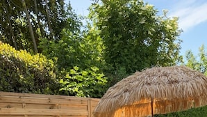 An outdoor patio with a wooden table and chairs, a thatched umbrella, and potted plants.