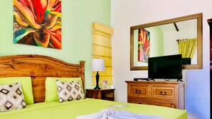Een net opgemaakt bed met een groene sprei, twee geborduurde kussens, een witte handdoek, een houten nachtkastje met een lamp, een kast met een televisie en een grote spiegel aan de muur.
