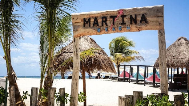 Un letrero de madera con la inscripción 'MARTINA BEACH', rodeado de palmeras y sombrillas de paja en una playa.