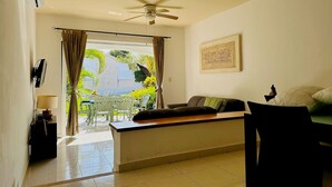 Una habitación con ventilador de techo, cama con almohadas, escritorio con lámpara y vista a follaje exterior.