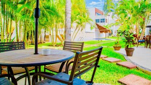A round table with chairs, a bench, and a potted plant in a courtyard with palm trees and a building in the background. - 22 Pax 2 Connected Pool Villas (Playa del Carmen)
