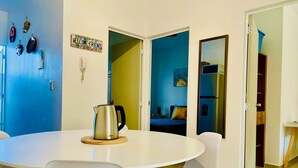 Un comedor con una mesa redonda, sillas blancas y un hervidor. Hay puertas que dan a otras habitaciones.