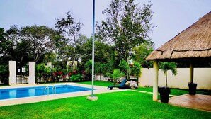 Una piscina con una estructura de techo de paja, rodeada por un césped cuidado y árboles.