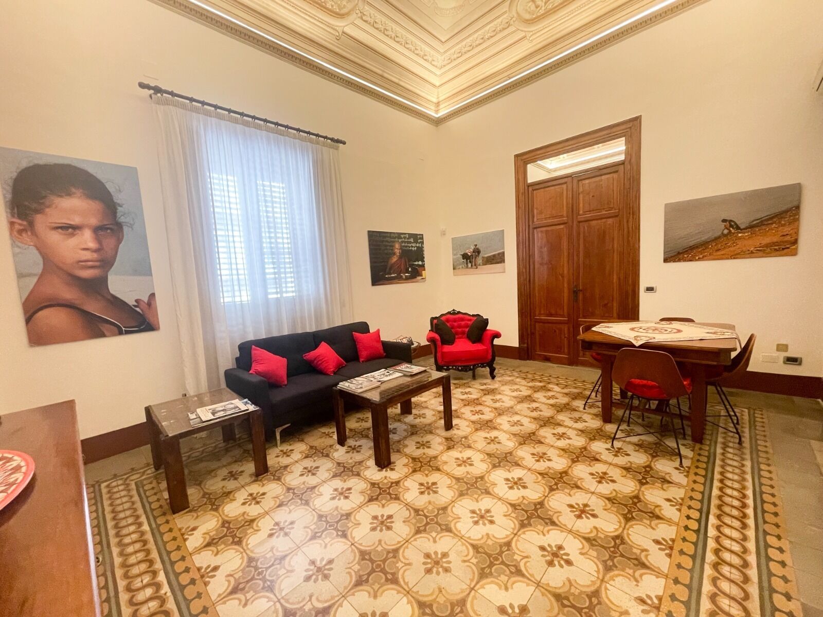 Ein Wohnzimmer mit einem gemusterten Boden, einem Sofa mit roten Kissen, einem roten Sessel, einem Holztisch und einem Esstisch mit Stühlen.