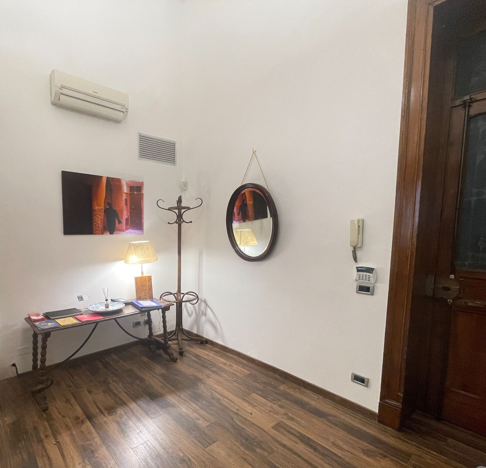 A room with a wooden floor, a desk with a lamp, a coat rack, a wall-mounted mirror, and a door.