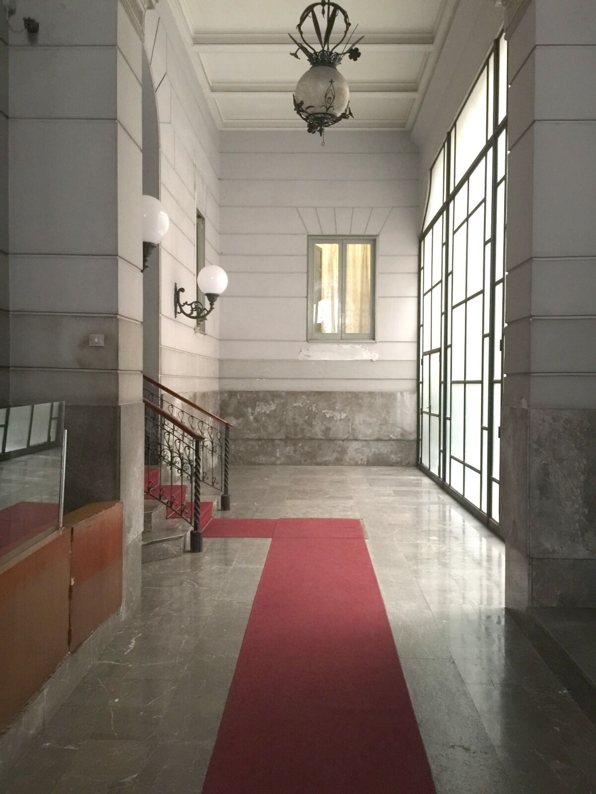 Um corredor com carpete vermelho, escada com grades de metal e uma janela grande com cortinas.