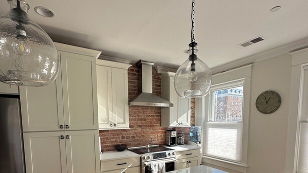 A modern kitchen with white cabinetry, a central island with a sink, and a brick wall. - Luxury townhome in Old town 2 blocks to King Street (Alexandria)