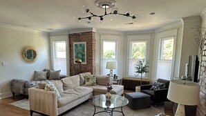 A spacious living room with a sectional sofa, a glass coffee table, and a dark armchair. There are large windows providing natural light and a view of greenery outside. - Luxury townhome in Old town 2 blocks to King Street (Alexandria)