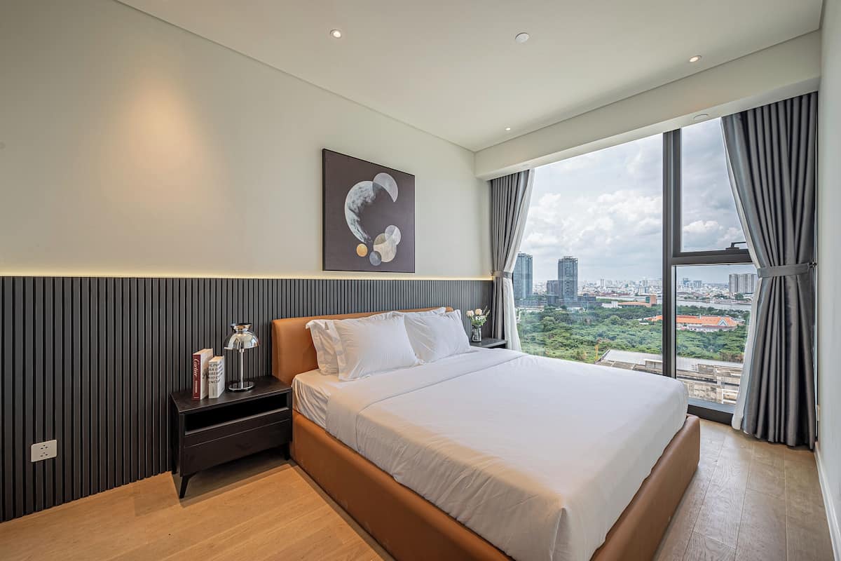 Quarto de hotel moderno com uma cama grande, uma mesa de cabeceira, uma obra de arte na parede e vista para a cidade através de janelas amplas.