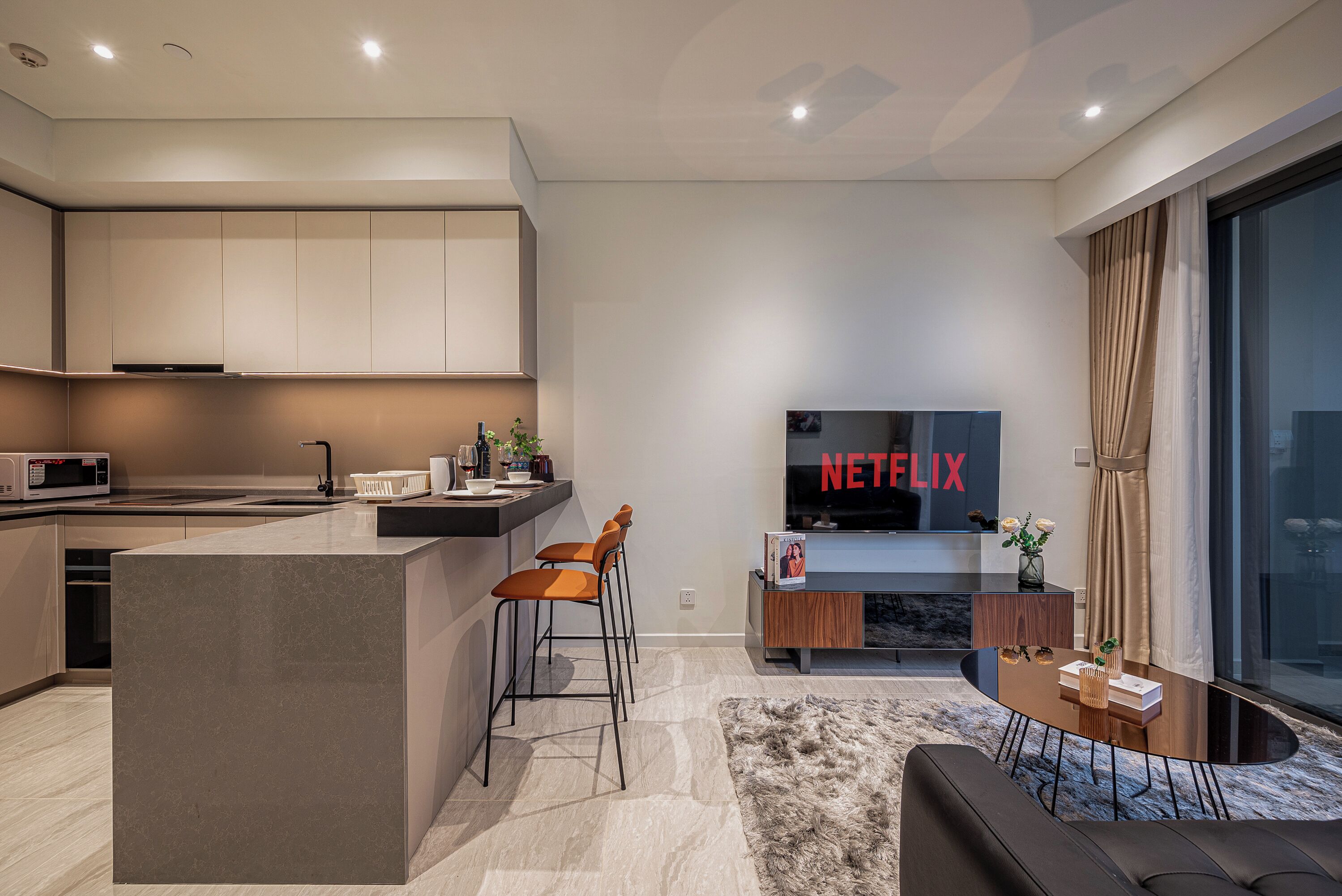 A modern kitchen with a central island, bar stools, a flat-screen TV, and a cozy seating area with a rug and sofa.