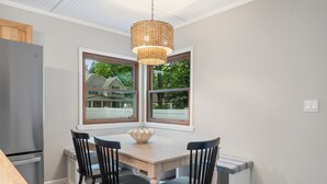 A dining area with a wooden table, four black chairs, a woven pendant light, and two windows offering a view of a house and trees. - Walk Downtown - Fall Colors, Wineries & Firepit (Boyne City)