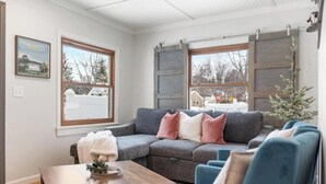 A cozy living room with a sectional sofa, a wooden coffee table, and a blue armchair. There are large windows with shutters, a framed picture on the wall, and a small Christmas tree. - Walk Downtown - Fall Colors, Wineries & Firepit (Boyne City)
