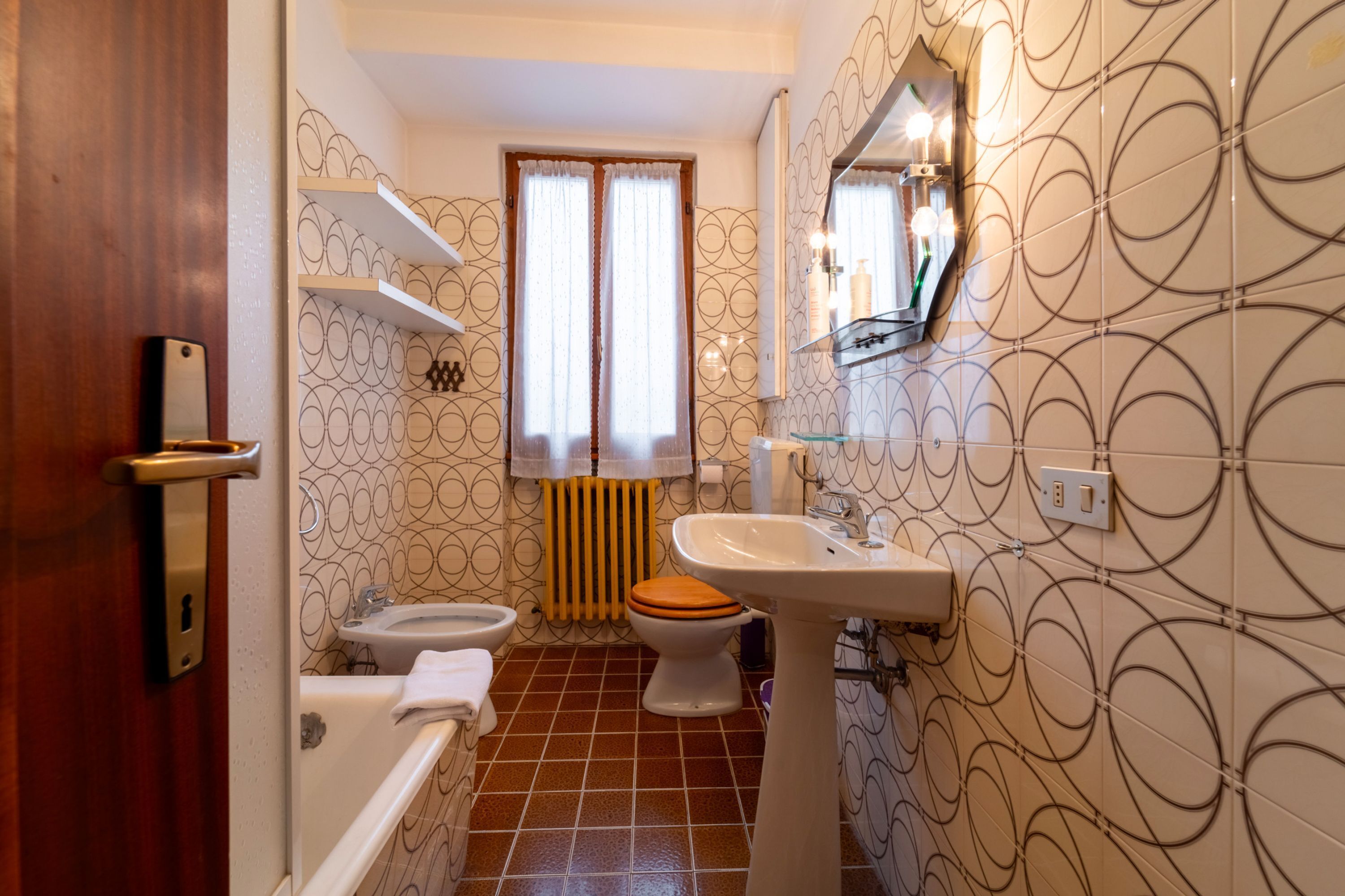 A bathroom with a toilet, sink, and bathtub. There is a window with curtains, a towel rack, and a mirror.