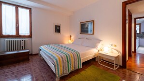 Un dormitorio con cama, mesita de noche, lámpara, radiador, banco y vistas a la cocina.