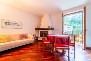 Un salon comprenant un canapé, une cheminée, une table à manger avec des chaises, et donnant sur un jardin grâce à une grande fenêtre.