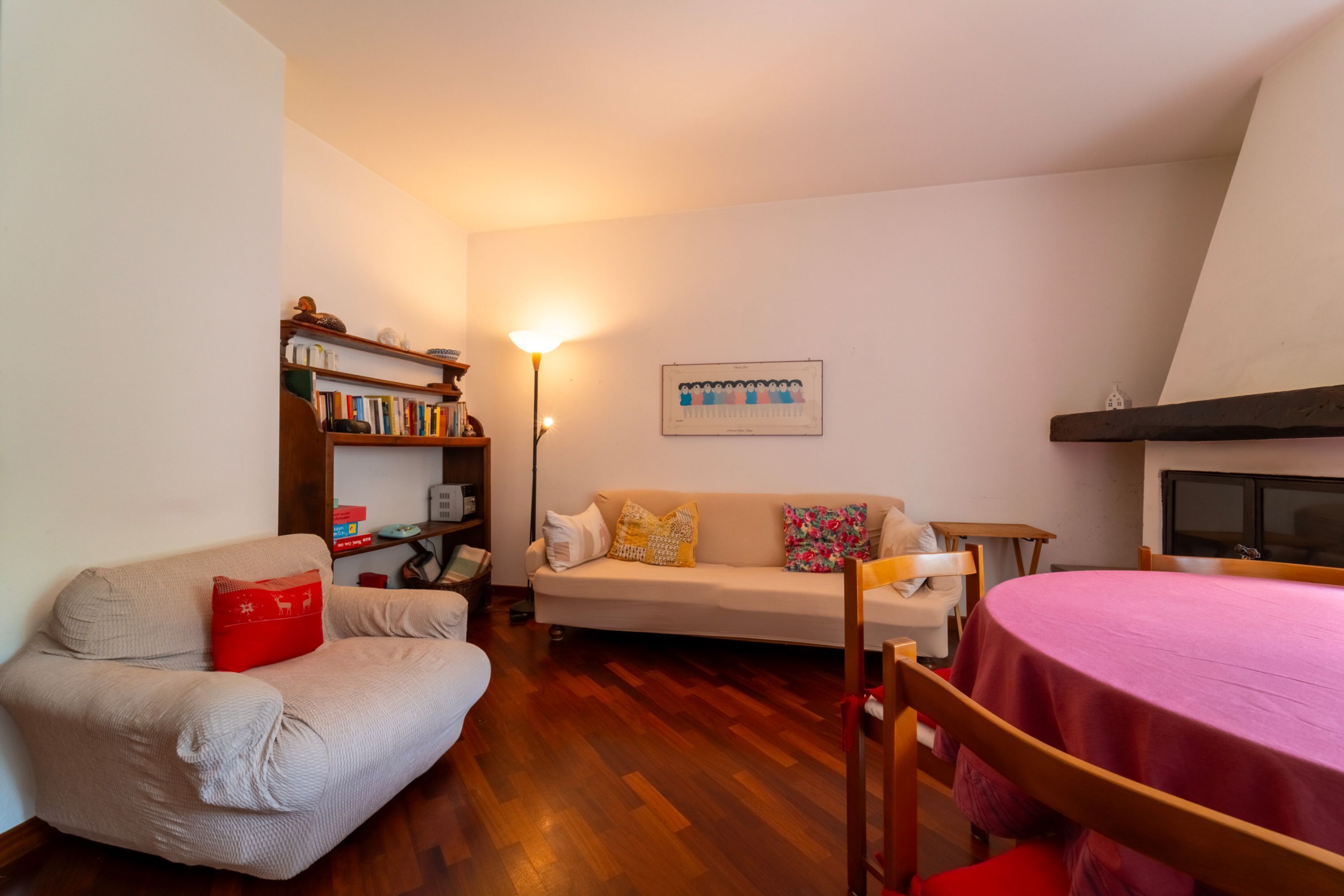 A cozy living room with a beige sofa, a red pillow, a wooden bookshelf, a pink table, and a framed picture on the wall.