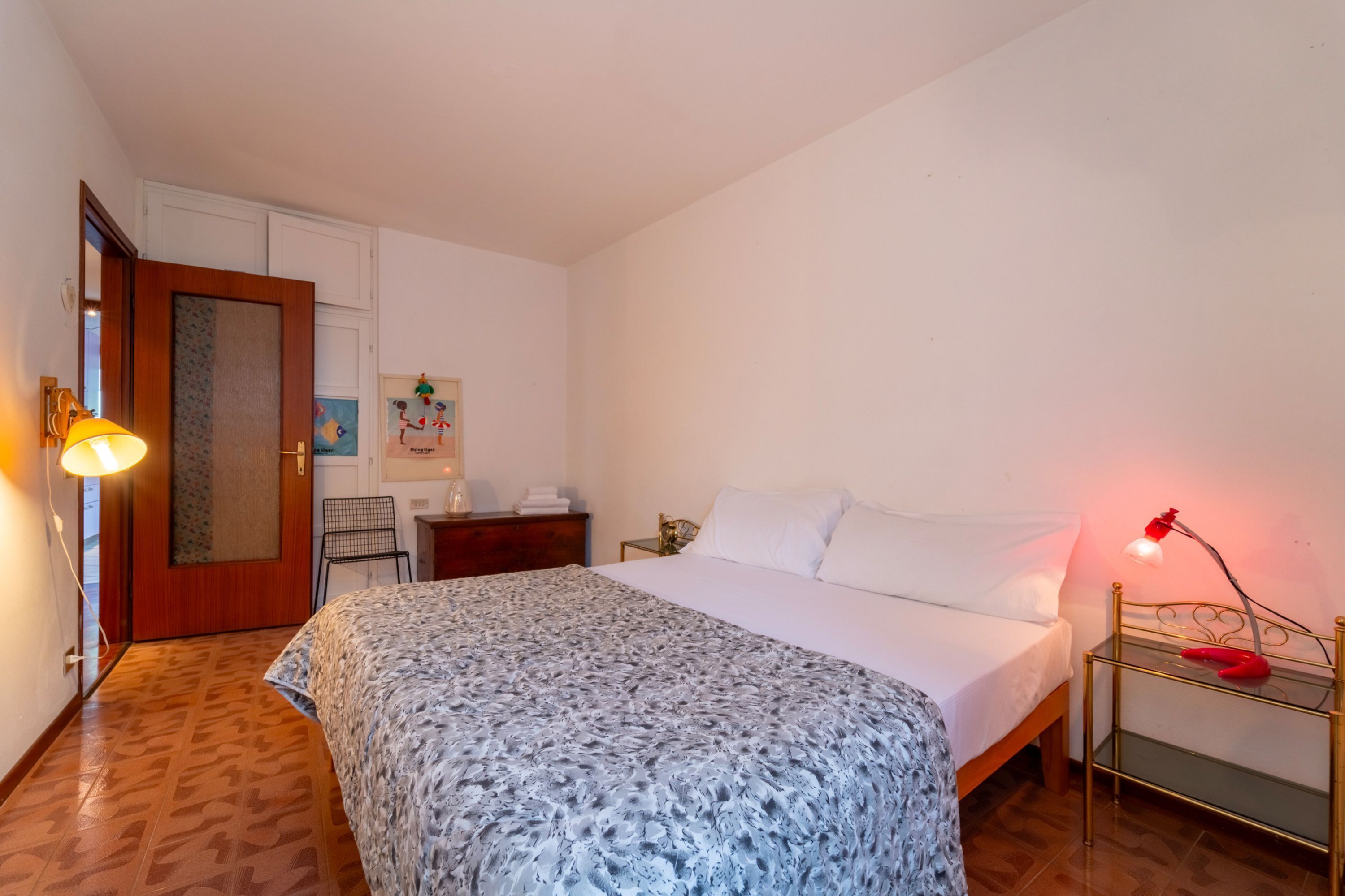 A bedroom with a bed, bedside table, lamp, and a door leading to another room.