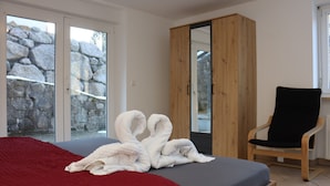 Un dormitorio con cama, una manta roja, un armario de madera, una silla negra y una pared de piedra visible a través de la puerta de cristal.