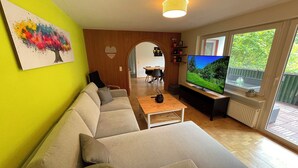 Sala de estar con sofá, mesa de centro y televisor. Hay un cuadro en la pared y una ventana con vistas a follaje.