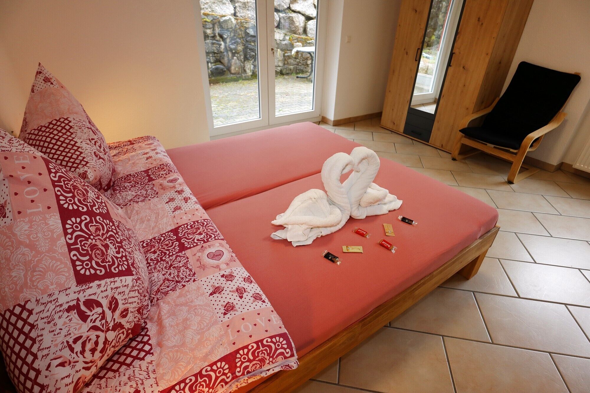A room with a bed, a wooden chair, and a window with a view of a stone wall and greenery.