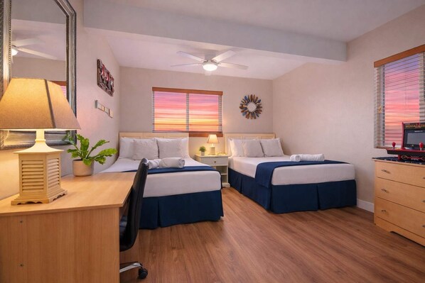 A hotel room with two beds, a desk with a lamp, a chair, a desk lamp, a potted plant, a ceiling fan, a window with blinds, and a wall clock.